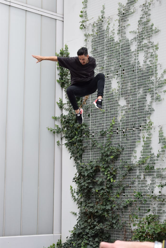 Man in black jumping against a wall with climbing plants, showcasing urban energy.